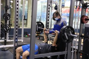 Un athlète couché sur un banc de musculation est accompagné par son entraîneur portant un masque qui s'assure de la sécurité du mouvement.