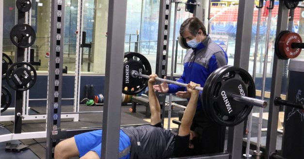 Un athlète couché sur un banc de musculation est accompagné par son entraîneur portant un masque qui s'assure de la sécurité du mouvement.